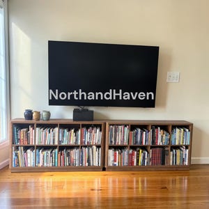 May include: A large flat-screen television with the text NorthandHaven displayed on the screen, mounted above two side-by-side wooden bookcases filled with numerous books and three small decorative ceramic vessels on the top surface.
