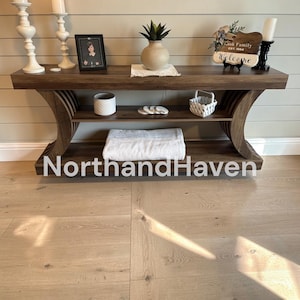 May include: A dark wood console table with a curved base and a lower shelf. The top surface holds two white candlesticks, a framed photo, a potted plant, and a sign reading "Rusk Family EST. 1994 Welcome". The lower shelf holds a white bowl and a folded white blanket.