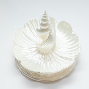 May include: A white ceramic dish featuring a central upright spiral seashell sculpture. The base of the dish is shaped like a flower with wide, curved petals radiating outward. The entire piece has a glossy, pearlescent finish that reflects light.