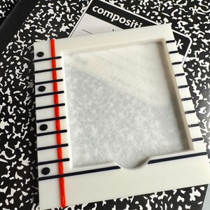 May include: A white 3D-printed frame designed to resemble a piece of notebook paper, featuring a vertical red line on the left side, black horizontal lines, and black dots along the margin. The frame has a central rectangular cutout.