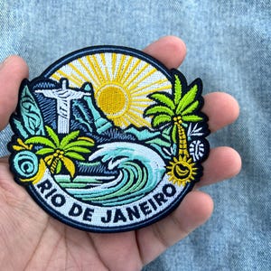Könnte beinhalten: Ein runder bestickter Aufnäher mit einer farbenfrohen Szene von Rio de Janeiro. Das Design zeigt die Christusstatue, eine leuchtend gelbe Sonne, grüne Palmen und eine große blaue Meereswelle, mit dem Schriftzug RIO DE JANEIRO am unteren Rand.