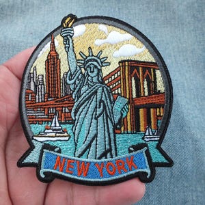 May include: A circular embroidered patch featuring the Statue of Liberty in the center, with the New York City skyline, the Brooklyn Bridge, and a boat in the background. The text NEW YORK is stitched in orange on a blue banner at the bottom.
