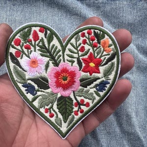 Può includere: Una toppa ricamata a forma di cuore con uno sfondo color crema e un vivace motivo floreale. Il ricamo presenta un grande fiore rosa centrale, un fiore rosa più chiaro, un fiore rosso, foglie verdi e piccole bacche di colore rosso.