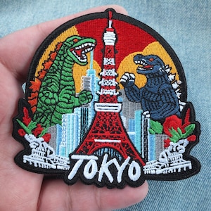 Peut inclure: Un écusson brodé circulaire représentant un monstre vert et un monstre bleu se faisant face devant la tour de Tokyo rouge et blanche, avec une ligne d'horizon urbaine en arrière-plan et le mot TOKYO brodé en blanc en bas.