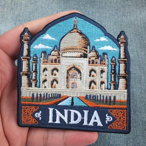 May include: An embroidered patch featuring the Taj Mahal in white and tan thread against a light blue sky with white clouds. The design includes four minarets, a central dome, and a dark blue base with the white text INDIA and orange accents.