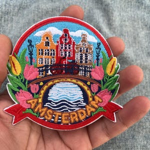 May include: A circular embroidered patch featuring a colorful scene of Amsterdam with three canal houses in orange, red, and brown, a bridge over water, and pink and yellow tulips. The word AMSTERDAM is stitched in gold letters on a red banner.
