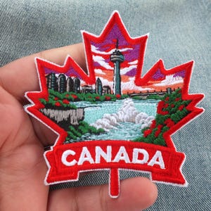 Puede incluir: Un parche bordado con forma de hoja de arce y borde rojo. El centro muestra un paisaje colorido de las cataratas del Niágara, la torre Skylon y un cielo al atardecer. La palabra CANADA está bordada en letras blancas sobre una cinta roja.