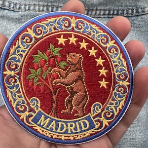 May include: A circular embroidered patch featuring a brown bear standing on its hind legs reaching for a strawberry tree on a red background. The design includes seven yellow stars, a blue border with gold scrollwork, and the text MADRID.