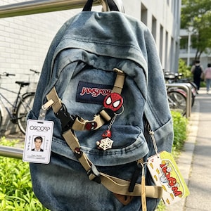 May include: A blue denim JanSport backpack featuring a beige nylon strap across the front, a red Spider-Man mask pin, a small character charm with a white web, an Oscorp Industries staff ID badge, and a rectangular yellow tag with red text.