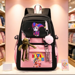 May include: A black and pink backpack featuring a cartoon character in a jester hat on the top pocket and the text "THE AMAZING DIGITAL CIRCUS" with character graphics on the bottom pocket. It includes a green frog keychain and a pink pom-pom.