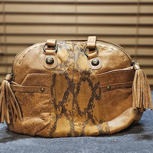 May include: A tan handbag featuring a central vertical panel with a brown snakeskin pattern. The bag includes two side tassels, antique brass-toned metal hardware, circular rings for the handles, and decorative stitching along the top edges.