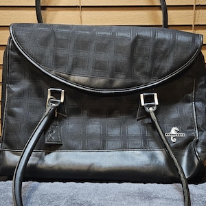 May include: A black handbag featuring a subtle grid pattern on the main body, a curved flap closure, and two rounded shoulder straps. The bag includes silver-toned hardware, a seahorse logo tag, and a metal plaque with the text ATLANTIC.