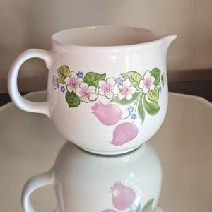 Peut inclure: Un pot à crème en céramique blanche doté d'un corps arrondi, d'une anse incurvée et d'un bec verseur. L'extérieur est orné d'un motif floral composé de fleurs roses ressemblant à des tulipes, de petites fleurs bleues et de feuilles vertes.