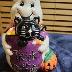 Op de afbeelding: Een keramieken koekjespot in de vorm van een witte geest die een zwarte kat en een pompoen vasthoudt. De geest houdt een paarse tas vast met de tekst Trick or Treat. Een geel meetlint toont een basisbreedte van ongeveer 24,1 cm.