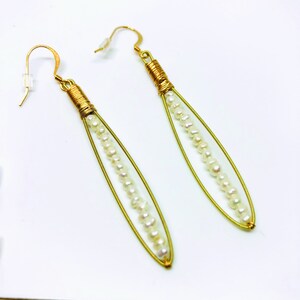 May include: A pair of gold earrings with a long, thin, oval shape. Each earring has a row of small white pearls strung along the center of the oval.