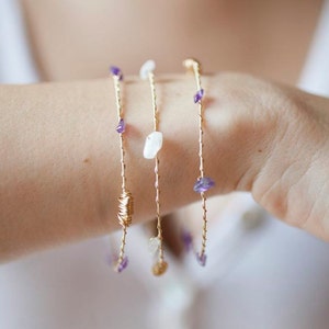 May include: Three gold bracelets with small purple and white stones. The bracelets are layered on a person's wrist.