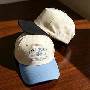 May include: Two baseball caps with cream-colored crowns. The front cap features a light blue brim and blue embroidery reading "LAST TOAST EST. 2025 On the Coast" with a seashell icon. The back cap has a black brim and the word "Destiny" embroidered.