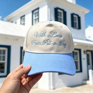 May include: A baseball cap with a cream-coloured crown and a light blue brim. The front of the crown features the embroidered text "With Love From Put-in-Bay" in a light blue cursive script. The cap has a structured five-panel design.