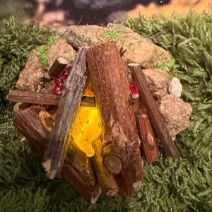 May include: A miniature campfire figurine constructed from small brown wooden twigs arranged in a conical shape over a glowing yellow translucent flame element, surrounded by a circular ring of textured grey stones and small red beads.