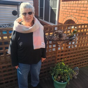 May include: A person wearing a black long-sleeved shirt, blue jeans, black trainers, and a light pink and white plaid scarf with a small rectangular label. The person has short grey hair and is wearing dark-tinted sunglasses.
