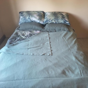 May include: A bed featuring a solid sage green blanket covering the mattress, topped with two solid sage green pillows and two additional pillows decorated with a green and white palm leaf pattern, all arranged against a plain light beige wall.