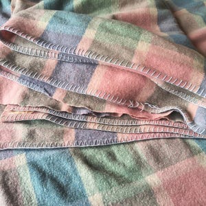 May include: A folded wool blanket featuring a plaid pattern in muted shades of pink, blue, green, and tan. The edges of the blanket are finished with a visible white blanket stitch, creating a decorative border along the folded fabric layers.
