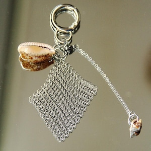 Puede incluir: Un llavero de metal plateado con una argolla circular, un panel de malla de cadena de forma cuadrada y dos pequeñas conchas de cauri moteadas en tonos beige y marrón, unidas por finas cadenas.
