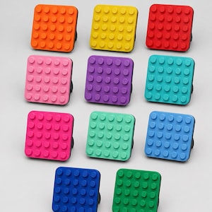 Può includere: Una collezione di undici pomelli quadrati per cassetti in vari colori solidi, tra cui arancione, giallo, rosso, rosa, viola, turchese, magenta, verde chiaro, azzurro, blu scuro e verde. Ogni pomello presenta una griglia di borchie circolari.