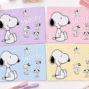 Pode incluir: Quatro cadernos retangulares nas cores pastel roxo, rosa, azul e amarelo. Cada capa apresenta uma grande ilustração do Snoopy, o texto SNOOPY e figuras menores do Snoopy em várias poses, incluindo uma fazendo o movimento de dança dab.