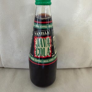 May include: A glass bottle of Java Cola Vanilla coffee-flavored soda with a green cap. The label features the text Java Cola and Vanilla in white and green lettering on a dark background, with a volume of 11 fl. oz. or 325 ml clearly printed.