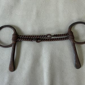 May include: A dark brown, rusted metal horse bit featuring a double-jointed mouthpiece with a twisted wire design. The bit includes two circular rings on each side, each connected to a downward-extending metal bar, showing a weathered texture.