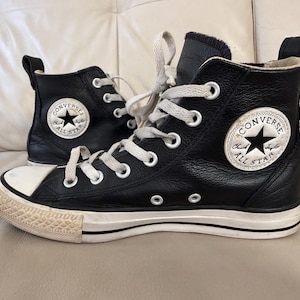 May include: A pair of black leather high-top sneakers with white laces, white rubber toe caps, and white rubber soles. Each shoe features a circular white patch on the side with the black text Converse All Star and a black star logo.