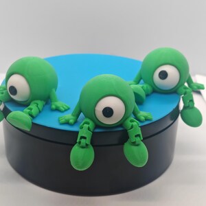 May include: Three small, green, spherical figurines with single large white eyes and black pupils. Each figure has articulated limbs and is positioned on a circular black and blue platform. The creatures have smooth, rounded bodies and jointed arms and legs.