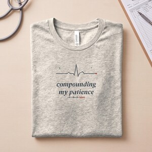 May include: A heather gray t-shirt featuring a black EKG heart rhythm line graphic with a small green plus sign and a red dot. Below the graphic, the text reads "compounding my patience" in a dark font, followed by a smaller EKG line with red accents.