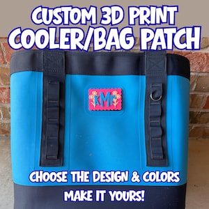 May include: A bright blue cooler bag with black trim and two vertical black webbing straps. A rectangular pink patch with scalloped edges is attached to the center, featuring the blue letters KMR and yellow floral designs on each side.