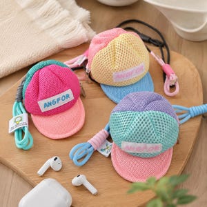 Può includere: Tre piccole pochette a rete a forma di cappellino con cerniere, ognuna con una toppa rettangolare bianca con la scritta ANGFOR in lettere blu o rosa. Le pochette sono rosa e turchese, giallo e rosa, o viola e turchese e includono cordini attaccati.