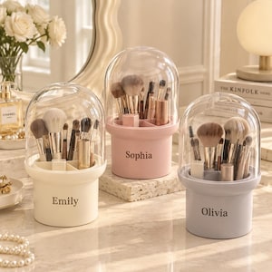 Puede incluir: Tres organizadores cilíndricos para brochas de maquillaje con tapas de cúpula transparentes. El recipiente izquierdo es crema con el nombre Emily, el central es rosa con el nombre Sophia y el derecho es gris claro con el nombre Olivia.