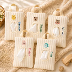 Puede incluir: Cinco fundas rectangulares para pañuelos de tela acolchada color crema con asas superiores y cremalleras. Cada funda presenta una cara de animal bordada sobre una abertura ovalada central y un nombre personalizado: Wrenly, Emma, Oliver, Daisy y Leo.