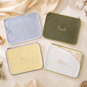 May include: Four rectangular zippered pouches in light blue, olive green, pale yellow, and white. Each pouch features a name embroidered in cursive script: Lily, Noah, Mia, and Ethan. The white pouch is partially open, revealing a light-colored garment.