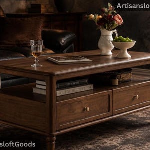Könnte beinhalten: Ein rechteckiger Couchtisch aus Holz mit dunkelbraunem Finish, ausgestattet mit zwei Schubladen mit kleinen runden Knöpfen und einer offenen Ablage. Auf der Oberfläche stehen ein Glas, ein Buch, ein weißer Krug mit Blumen und eine Schale mit grünen Trauben.