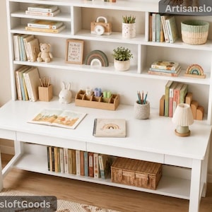 Puede incluir: Un escritorio de madera blanca con una estantería integrada que contiene libros, un pequeño oso de peluche, una cámara de juguete de madera, una planta en maceta, una cesta tejida y una lámpara. La superficie del escritorio tiene un libro y un cuaderno.