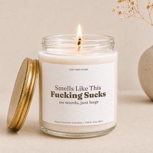 May include: A clear glass jar candle with a gold metal lid, filled with white wax and a lit wick. The white label features the text: COZY WAX STUDIO, Smells Like This Fucking Sucks, no words, just hugs, Hand Poured Candles | 100% Soy Wax.