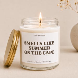 May include: A lit soy wax candle in a clear glass jar with a gold metal lid. The white label features the text COZY WAX STUDIO, SMELLS LIKE SUMMER ON THE CAPE, and Hand Poured Candles | 100% Soy Wax in dark brown lettering against a cream background.