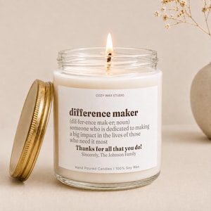 May include: A clear glass jar candle with a lit wick and a gold-colored metal lid. The white label features the text: COZY WAX STUDIO, difference maker (dif·fer·ence mak·er; noun) someone who is dedicated to making a big impact in the lives of those who need it most. Thanks for all that you do! Sincerely, The Johnson Family. Hand Poured Candles | 100% Soy Wax.