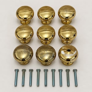 May include: A set of nine round, polished brass cabinet knobs arranged in three rows of three, accompanied by nine matching silver-toned metal mounting screws. The knobs feature a smooth, reflective surface with some visible signs of wear.
