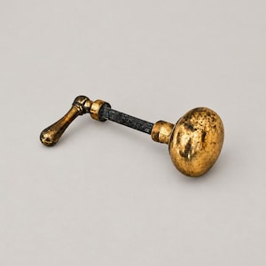 May include: A brass door knob with a distressed, aged gold finish and a dark, weathered patina. The hardware includes a round knob attached to a central metal spindle, which connects to a small, curved lever handle on the opposite end.