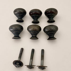 May include: A collection of six round, dark metallic cabinet knobs with a weathered, aged finish. The knobs feature a smooth, domed top and a tapered base. Three separate threaded metal bolts with attached washers are positioned below the knobs.