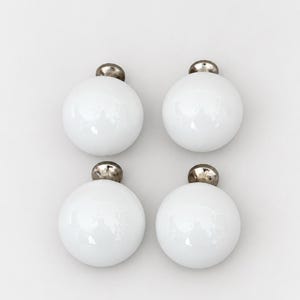 May include: A set of four spherical white knobs with a smooth, glossy finish. Each knob features a small, metallic silver-toned circular base at the top, providing a clean and simple design suitable for furniture hardware or cabinet drawer pulls.