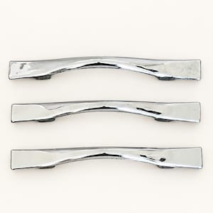 May include: Three identical cabinet pulls with a polished chrome finish. Each handle features a smooth, slightly arched design with flat, rectangular ends and two mounting points on the underside, arranged horizontally in a vertical stack.
