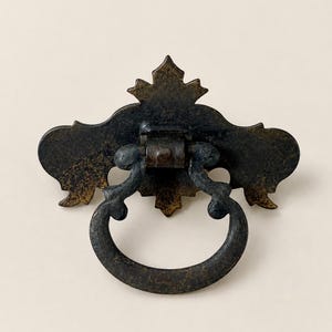 May include: A dark bronze-toned metal drawer pull featuring an ornate, decorative backplate with pointed edges and a circular, hinged drop handle. The hardware displays a weathered, textured finish with visible signs of aged patina throughout.